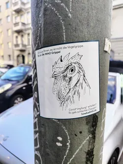 A square sticker affixed to a pole shows a hand-drawn illustration of a rooster's head. Above the drawing, the text reads 'Ich sage Ihnen, es ist nicht die Vogelgrippe. Es ist die WHO Grippe!' Below the image, it says 'Covid "Impfung" erhalten? Es gab schlechte Chargen!' and 'howbadinfo.de'.
