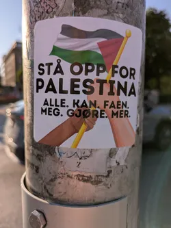 The sticker prominently displays the Palestinian flag waving at the top. Below the flag, two hands, one with a darker skin tone and the other lighter, are shown grasping the yellow flagpole together, symbolizing unity and shared effort. The text on the sticker is in Norwegian, set in a clear, bold, dark font against a white background. The sticker is affixed to a grey metal pole, which shows signs of wear and some faint markings from other removed adhesives. The background is blurred, suggesting an urban outdoor setting.