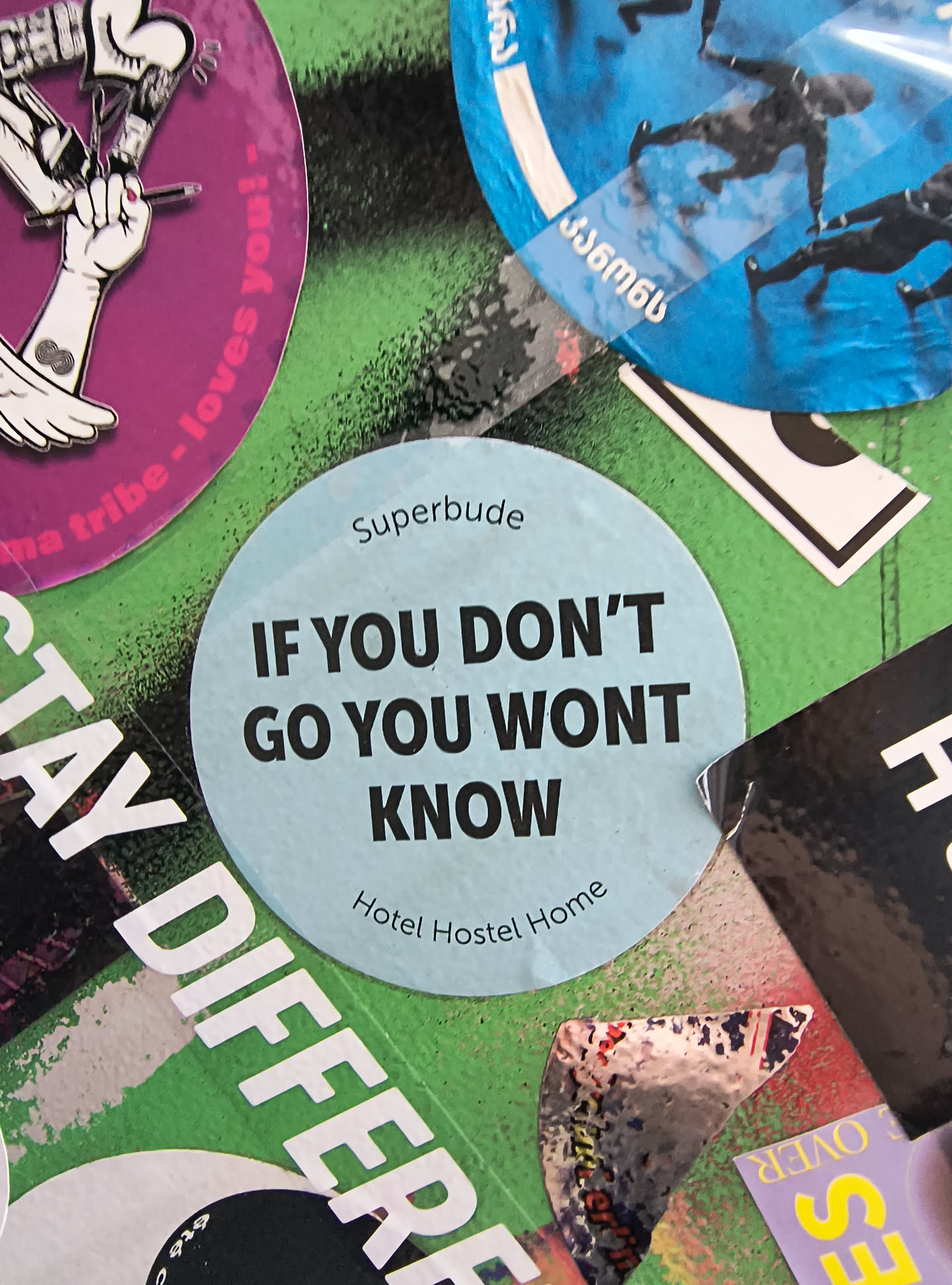 A circular, light blue sticker with bold black text. The top of the sticker features the word 'Superbude' in a smaller font. The main text reads 'IF YOU DON'T GO YOU WONT KNOW' in large, uppercase letters. Below this, in a smaller, curved font, it says 'Hotel Hostel Home'. The sticker appears to be placed on a surface covered with other stickers and graffiti.