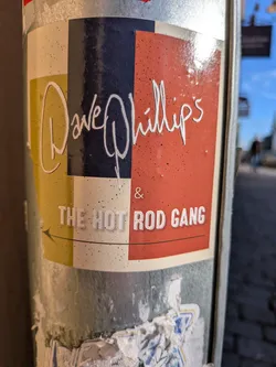 The sticker features a rectangular color-blocked composition divided into columns and sections of muted yellow, dark navy, cream, and reddish-orange. Across the top portion, the name 'Dave Phillips' is written in a stylized, flowing white script font. Below this, separated by an ampersand, is the text 'THE HOT ROD GANG' in bold, white, stylized block capitals. Underneath the band name is a thin line ending with a left-pointing arrow.