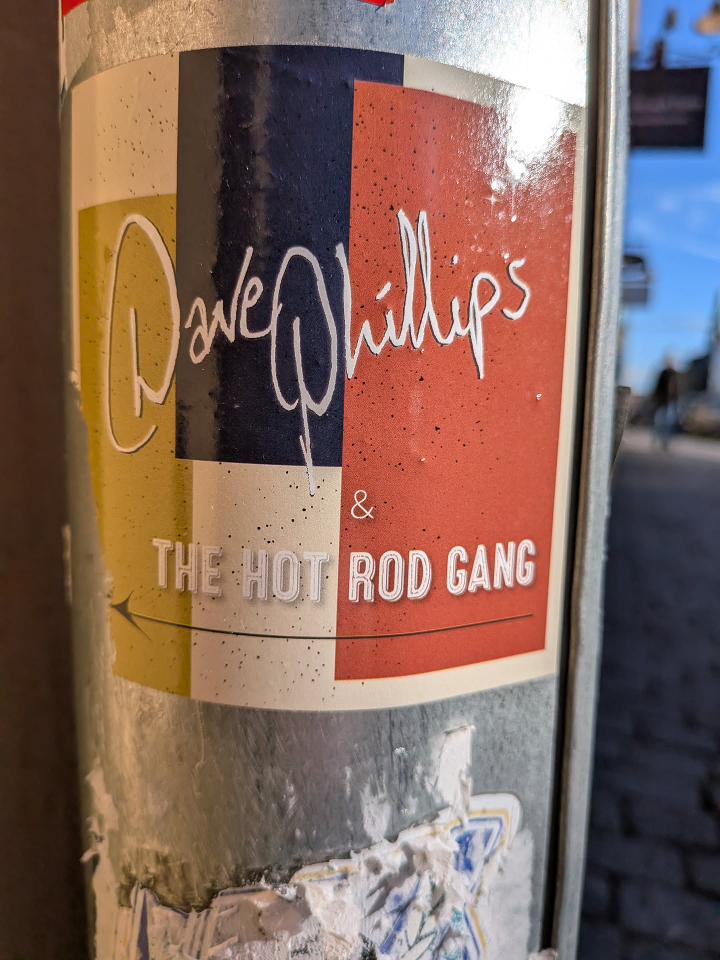 The sticker features a rectangular color-blocked composition divided into columns and sections of muted yellow, dark navy, cream, and reddish-orange. Across the top portion, the name 'Dave Phillips' is written in a stylized, flowing white script font. Below this, separated by an ampersand, is the text 'THE HOT ROD GANG' in bold, white, stylized block capitals. Underneath the band name is a thin line ending with a left-pointing arrow.