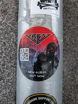 A circular sticker featuring a dark, futuristic, and menacing character with glowing red eyes and a spiked outfit. The character stands against a backdrop of a dark, dystopian city skyline with purple and blue hues. Above the character, on a vibrant red background, is the stylized black and silver logo 'PRIEZ'. To the left of the character, a white QR code is prominently displayed. Below the character, white text reads 'NEW ALBUM OUT NOW'. The overall aesthetic is dark, aggressive, and reminiscent of heavy metal or cyberpunk themes.