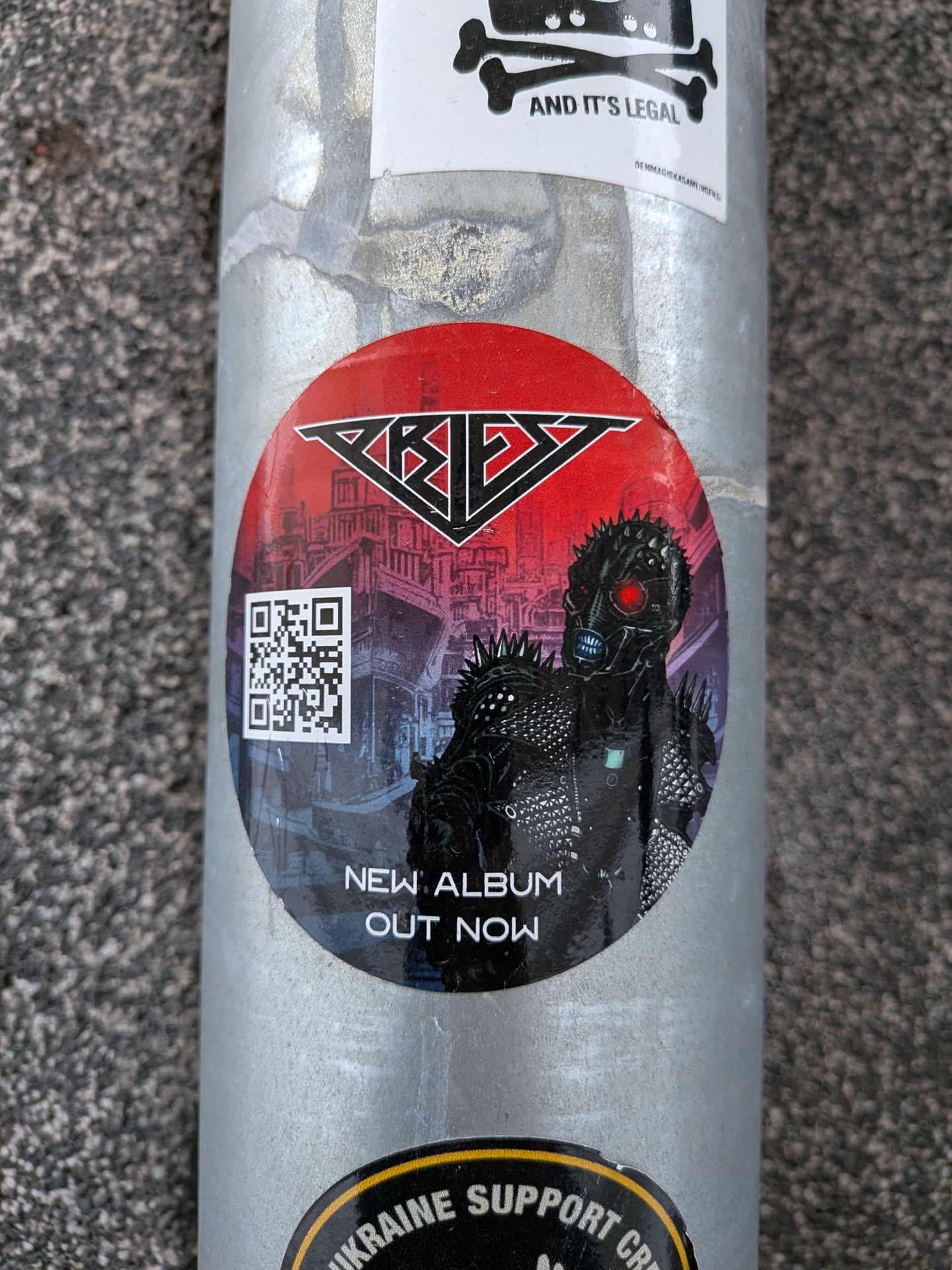 A circular sticker featuring a dark, futuristic, and menacing character with glowing red eyes and a spiked outfit. The character stands against a backdrop of a dark, dystopian city skyline with purple and blue hues. Above the character, on a vibrant red background, is the stylized black and silver logo 'PRIEZ'. To the left of the character, a white QR code is prominently displayed. Below the character, white text reads 'NEW ALBUM OUT NOW'. The overall aesthetic is dark, aggressive, and reminiscent of heavy metal or cyberpunk themes.