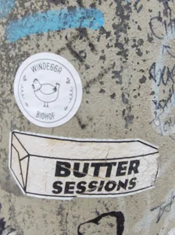 A sticker featuring the words "Butter Sessions" in a bold, sans-serif font.  The sticker is shaped like a rectangular block, reminiscent of a stick of butter. There's a smaller circular sticker above it with a chicken and text that reads "WINDE66A" and "BIOHOF".