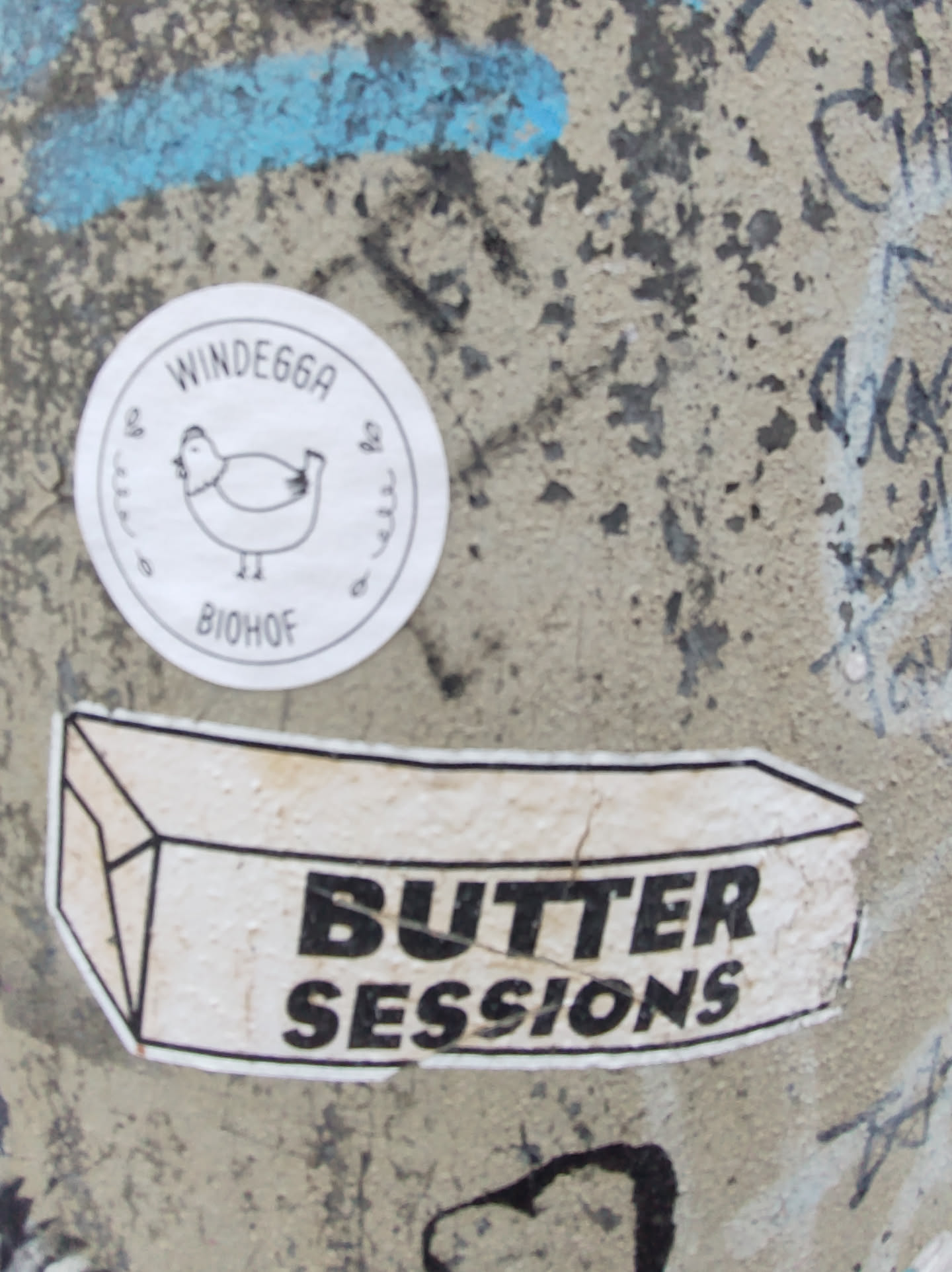 A sticker featuring the words "Butter Sessions" in a bold, sans-serif font.  The sticker is shaped like a rectangular block, reminiscent of a stick of butter. There's a smaller circular sticker above it with a chicken and text that reads "WINDE66A" and "BIOHOF".