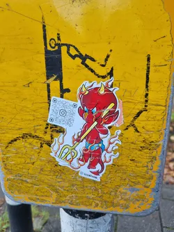 A sticker featuring a cartoon devil with flames, holding a pitchfork. The sticker has a QR code and the artist's signature. 