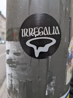The sticker is circular with a black background. In the center, the word 'IRREGALIA' is written in a stylized, white, serif font. Below the text is a white, abstract, flowing shape that resembles a stylized 'R' or a dripping form. The overall design is minimalist and graphic.