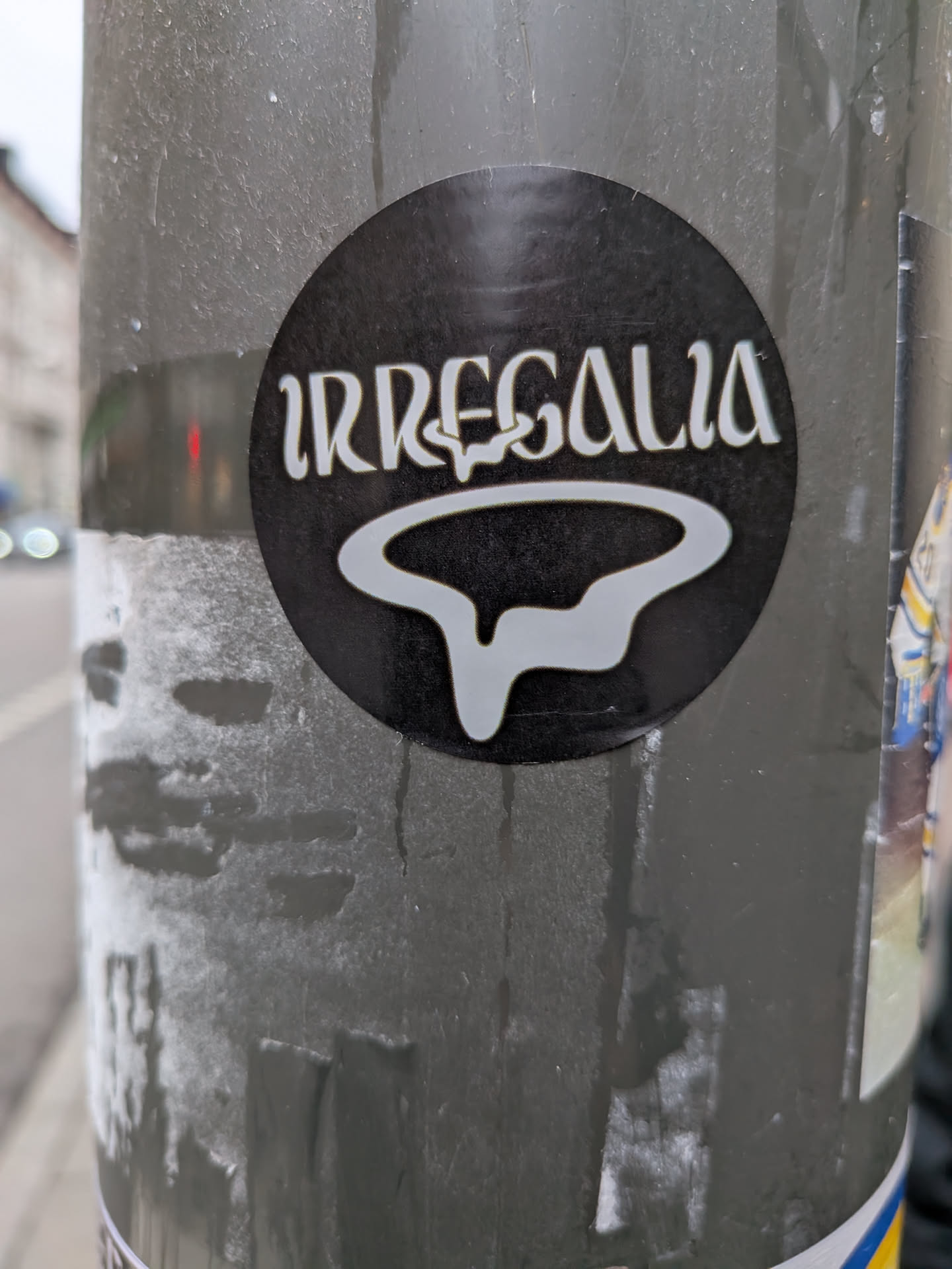 The sticker is circular with a black background. In the center, the word 'IRREGALIA' is written in a stylized, white, serif font. Below the text is a white, abstract, flowing shape that resembles a stylized 'R' or a dripping form. The overall design is minimalist and graphic.