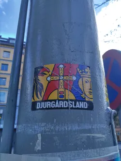 A sticker featuring a stylized coat of arms with a bird, cross, and crowned figure. The text 'DJURGÅRDSLAND' is visible at the bottom.