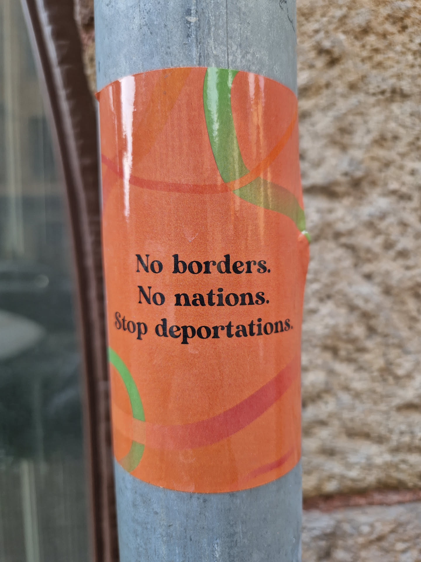 An orange sticker with green and red abstract shapes. The text on the sticker is in black and says: No borders. No nations. Stop deportations.