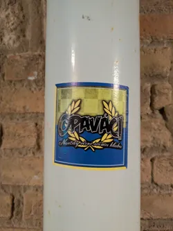 A rectangular sticker with gold, blue and black design, featuring the word "Opavači" between two laurel wreaths, and the text "Navždy věrni svému klubu" below, attached to a white pipe, which is in front of a brick wall.