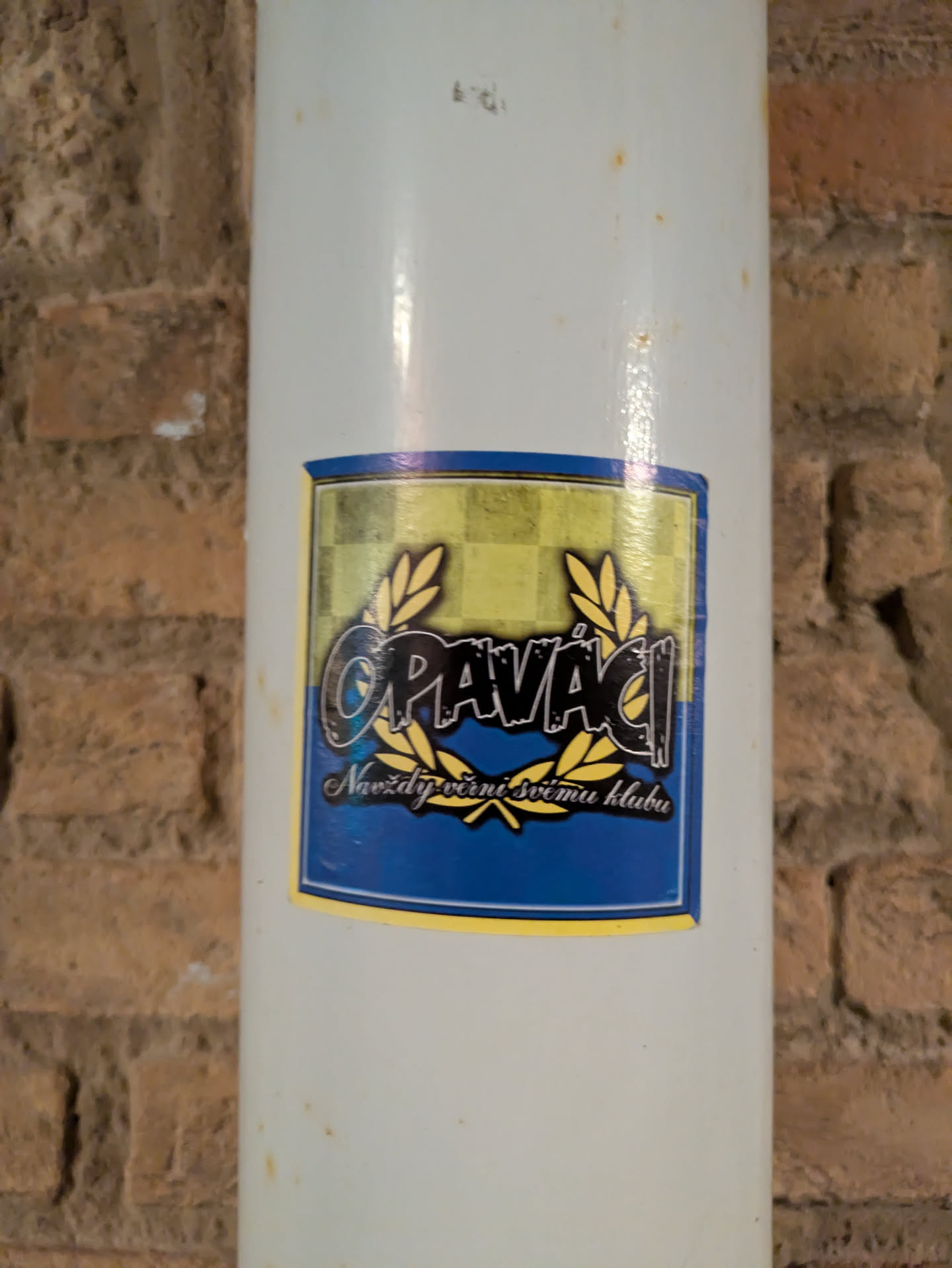 A rectangular sticker with gold, blue and black design, featuring the word "Opavači" between two laurel wreaths, and the text "Navždy věrni svému klubu" below, attached to a white pipe, which is in front of a brick wall.