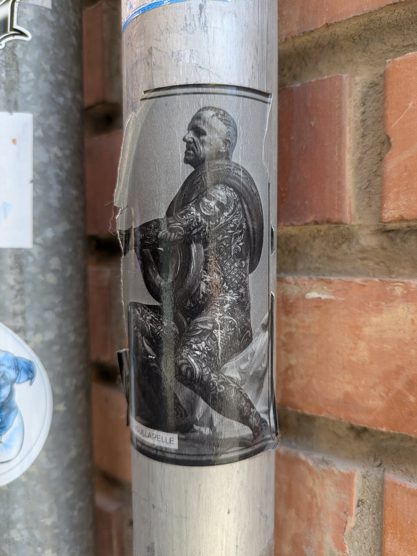 A sticker depicts a man covered in tattoos, sitting and holding something cylindrical. The sticker is peeling in places and affixed to a pole.