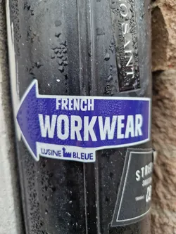 A purple and white sticker with an arrow pointing to the left. The text on the sticker says "FRENCH WORKWEAR" and below it in smaller font "LUSINE BLEUE". The sticker is affixed to a dark-colored cylindrical object that appears to be outdoors and slightly wet.