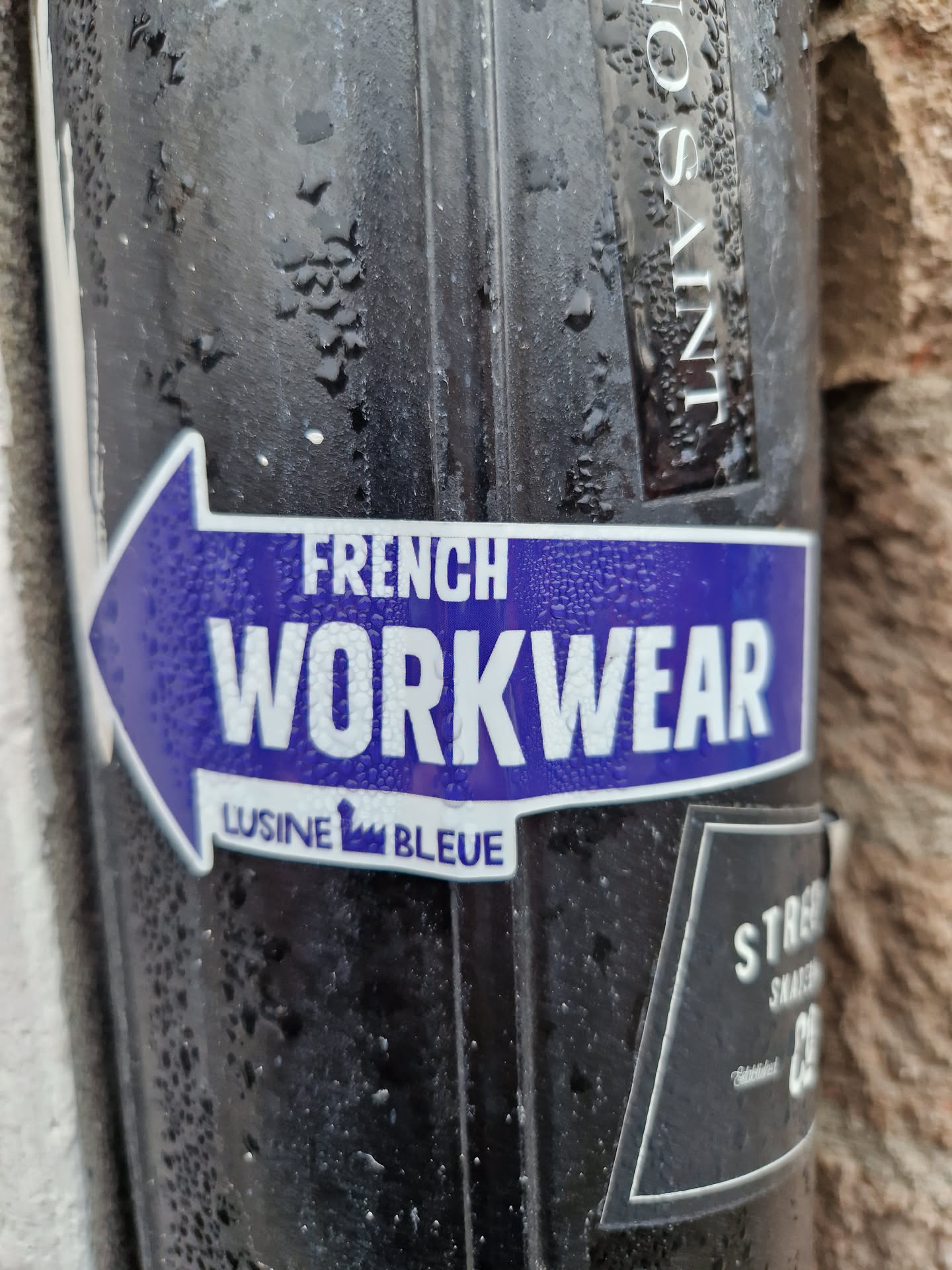 A purple and white sticker with an arrow pointing to the left. The text on the sticker says "FRENCH WORKWEAR" and below it in smaller font "LUSINE BLEUE". The sticker is affixed to a dark-colored cylindrical object that appears to be outdoors and slightly wet.