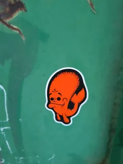 The sticker features a cartoonish character with an orange body and a black outline. The character has a large, round head with two black dots for eyes and a wide, smiling mouth with visible teeth. Its body appears somewhat amorphous, with two stubby arms and a tail-like appendage. The top of its head is textured with black stippling. The sticker has a white border.