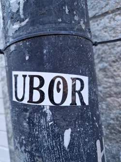 A close-up shot of a black and white sticker with the word "UBOR" printed on it. The sticker is affixed to a dark gray pole that shows signs of wear and tear.