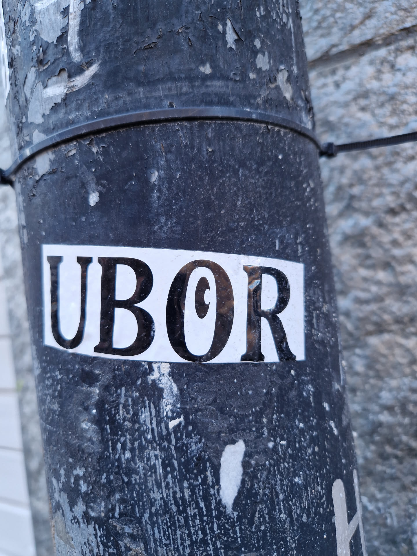 A close-up shot of a black and white sticker with the word "UBOR" printed on it. The sticker is affixed to a dark gray pole that shows signs of wear and tear.
