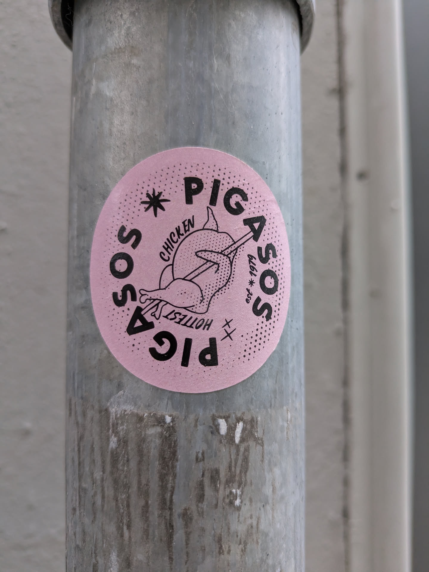 This is a round, pink sticker with black typography and an illustration, affixed to a grey metal pole. The central graphic depicts a stylized chicken or rooster with an arrow piercing through it. The text "PIG" is prominently displayed at the top and inverted at the bottom of the circle. Along the left edge, the word "SOS" is written curving upwards, and along the right edge, "ASOS" curves downwards, accompanied by "est * 1979". Smaller text near the chicken reads "CHICKEN" and "HOTTEST". The pink background has a subtle dotted texture, particularly around the edges.
