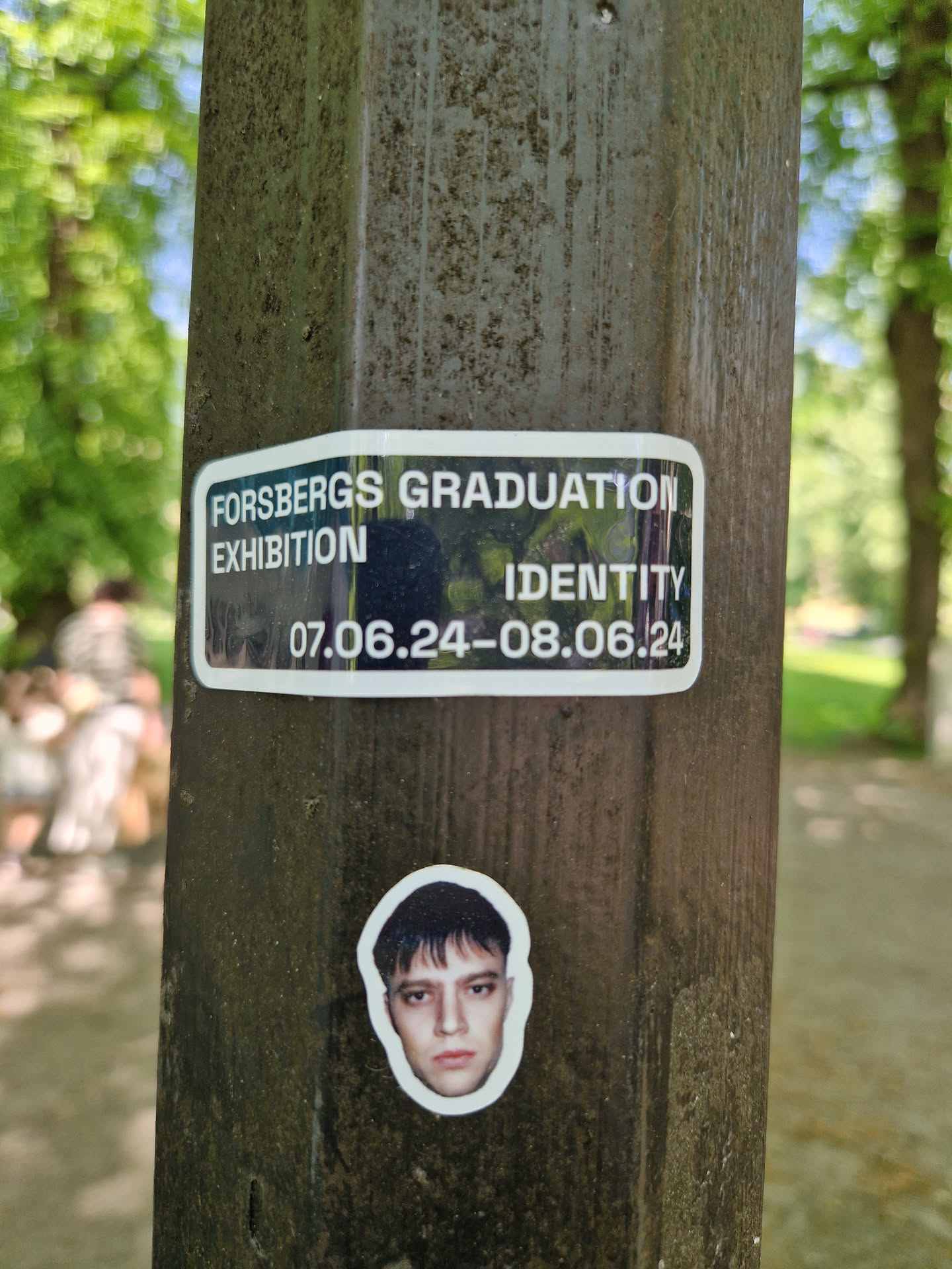 A sticker featuring the text "Forsbergs Graduation Exhibition Identity" and dates 07.06.24-08.06.24, along with a headshot of a person. The sticker is affixed to a dark-colored pole outdoors.