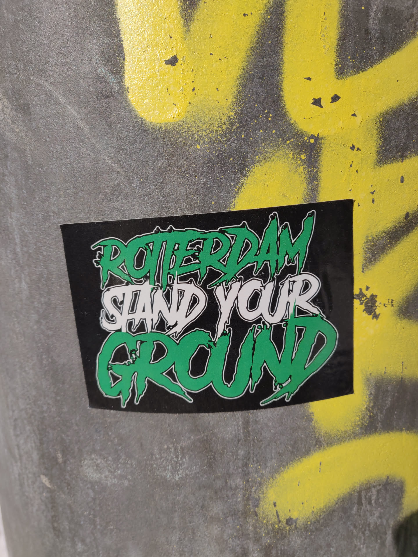 A black sticker with green and white text on a gray surface with yellow graffiti. The text says "Rotterdam Stand Your Ground".