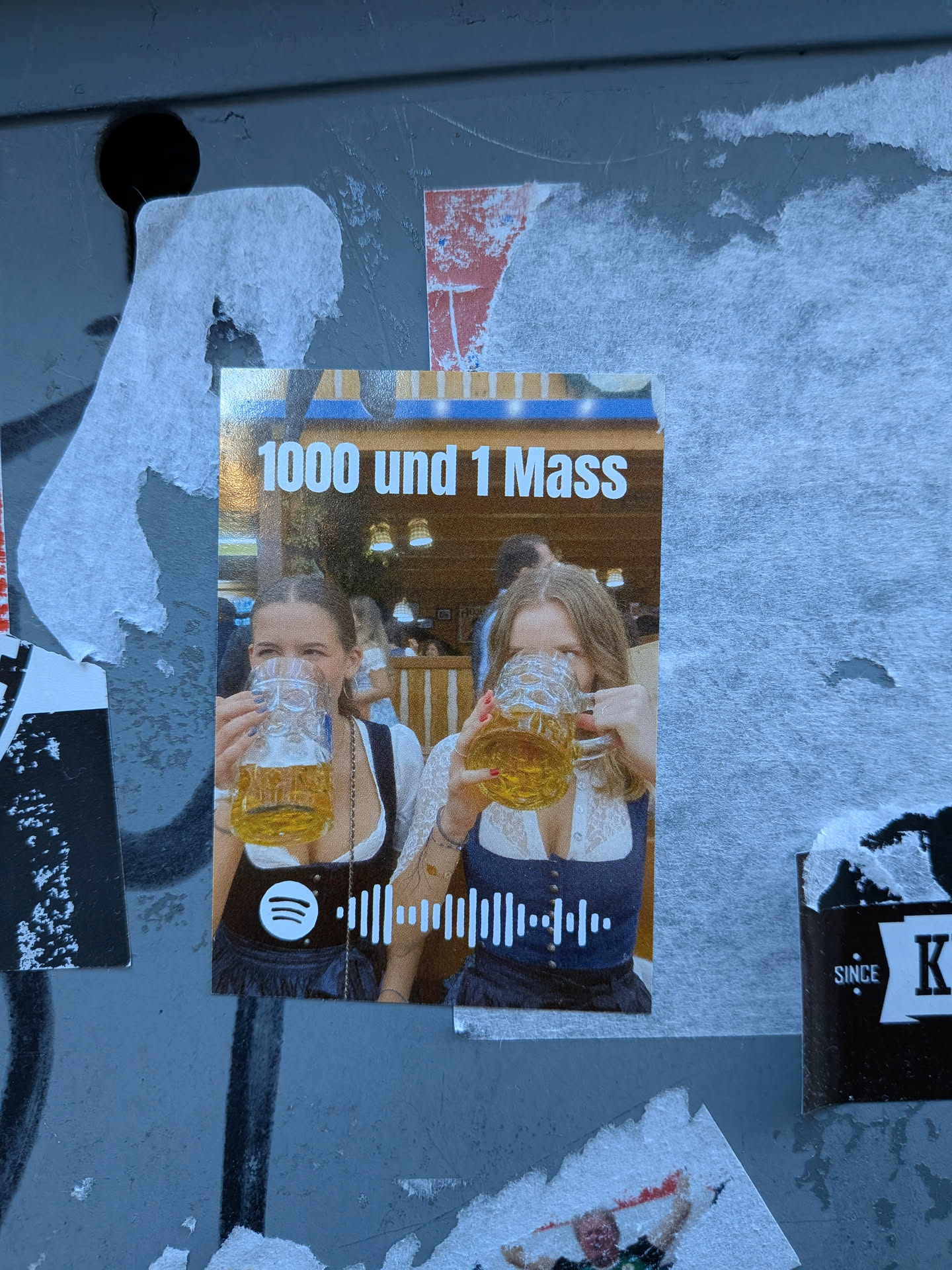 A rectangular sticker featuring a vibrant photograph of two young women, each holding a large glass beer mug (Masskrug) filled with golden beer. They are smiling and looking towards the viewer, dressed in traditional Bavarian dirndls (white blouses with lace details, dark bodices, and skirts). The background is a blurry, warm-toned indoor setting, possibly a beer hall. At the top, white text reads '1000 und 1 Mass'. At the bottom, a white Spotify logo is visible on the left, followed by a white Spotify audio wave code on the right. The sticker is adhered to a grey, textured surface, with some visible wear and tear around its edges.