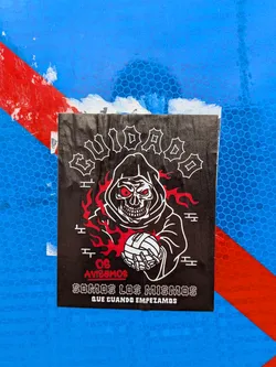 The sticker features a black background with a stylized grim reaper figure in white outline. The reaper has a skull face with glowing red eyes and is wearing a hooded robe. Flames in red and white emanate from the sides of the reaper's head. The reaper is holding a white soccer ball in its skeletal hands. Above the reaper, the word 'GUIADO' is written in a gothic, outlined font. Below the reaper, the text 'OS AVISAMOS' is in red, followed by 'SOMOS LOS MISMOS' and 'QUE CUANDO EMPEZAMOS' in a similar gothic font to 'GUIADO'. The sticker appears to be slightly worn at the edges.