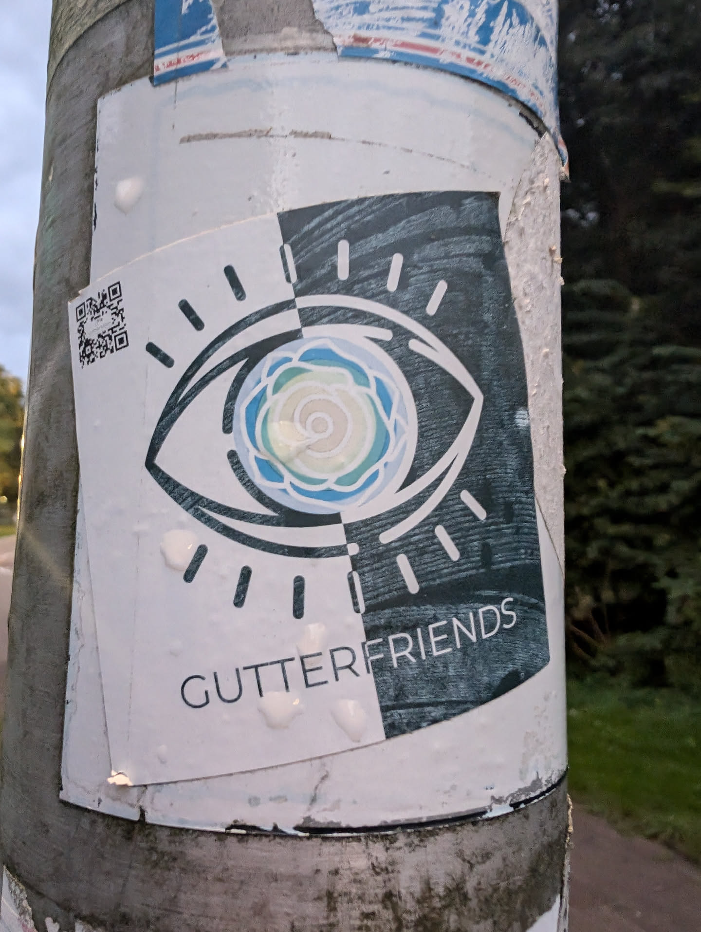 A sticker featuring a stylized eye graphic, split down the middle with a contrasting color scheme. One side is white with a light blue and green flower-like design within the eye, while the other is dark gray with a wood-grain texture. The text 'Gutterfriends' is visible at the bottom. A QR code is also present on the sticker.