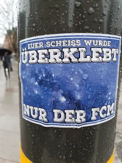 A blue and white sticker with German text on a dark pole. The text says "Euer Scheiss wurde überklebt" which translates to "Your shit has been pasted over." and "Nur der FCM", which seems to be an abbreviation or a name.