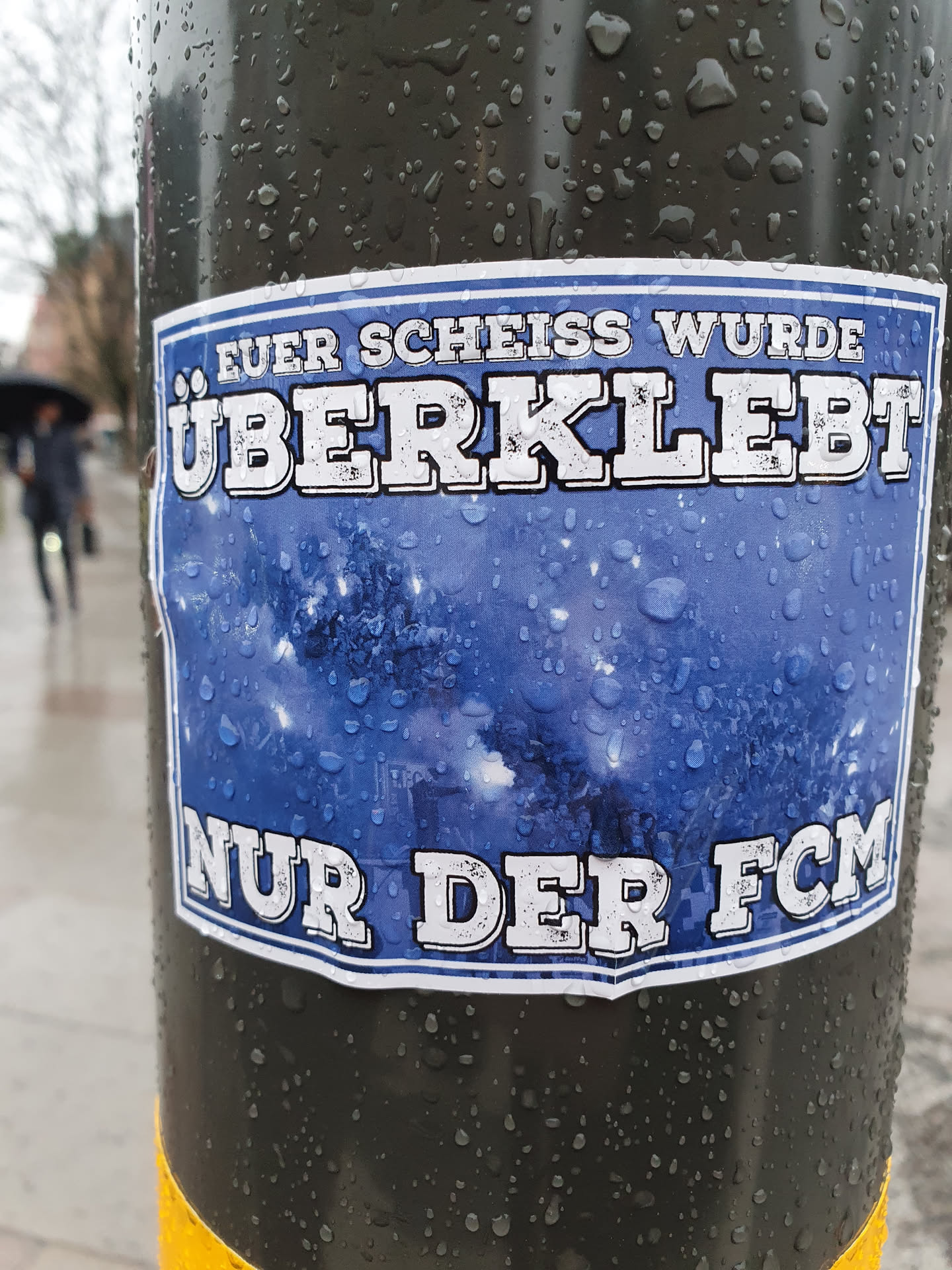 A blue and white sticker with German text on a dark pole. The text says "Euer Scheiss wurde überklebt" which translates to "Your shit has been pasted over." and "Nur der FCM", which seems to be an abbreviation or a name.