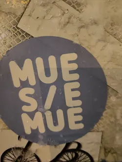 Round sticker with the text "MUES/MUE" written vertically. The sticker is light purple and the text is white. The sticker appears to be placed on a wall outside.