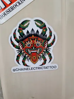 A sticker featuring a traditional style tattoo of a crab with a demonic face. The crab is predominantly red and green with black detailing. The face is stylized with exaggerated features and has a fierce expression.  The artist's Instagram handle is visible on the sticker. 