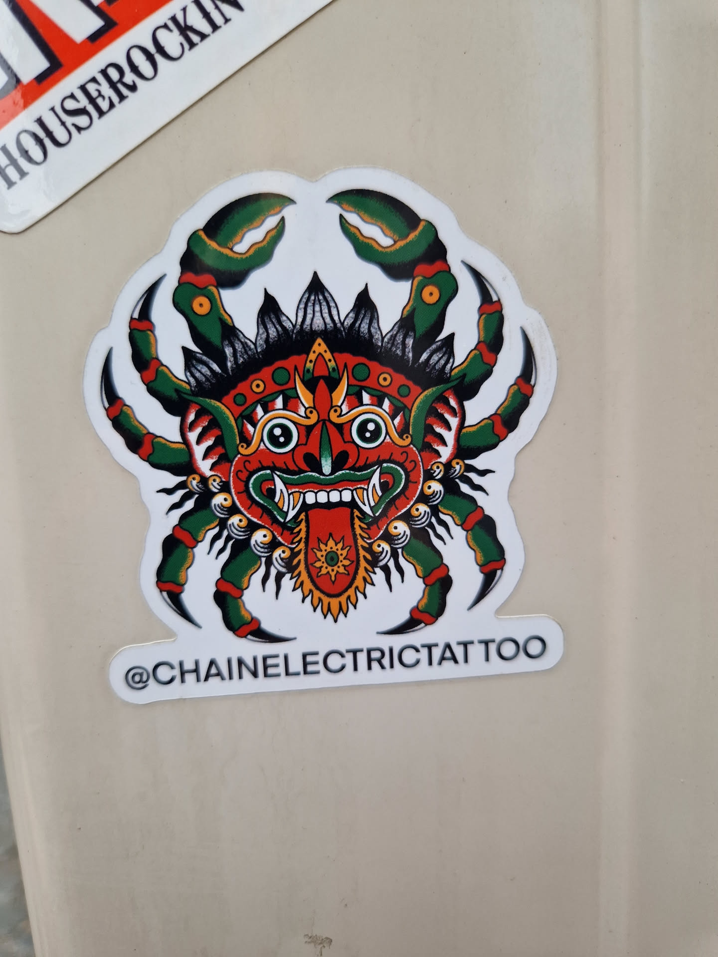 A sticker featuring a traditional style tattoo of a crab with a demonic face. The crab is predominantly red and green with black detailing. The face is stylized with exaggerated features and has a fierce expression.  The artist's Instagram handle is visible on the sticker. 