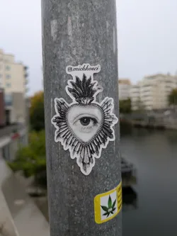 A die-cut black-and-white sticker placed vertically on a galvanized metal pole. The artwork features a sacred heart motif adorned with radiating spikes or rays and topped with flames. In the center of the heart is a realistically drawn human eye executed in a fine stippling and dotwork style. At the top of the sticker, an Instagram logo precedes the cursive handle '@miekkosci'.