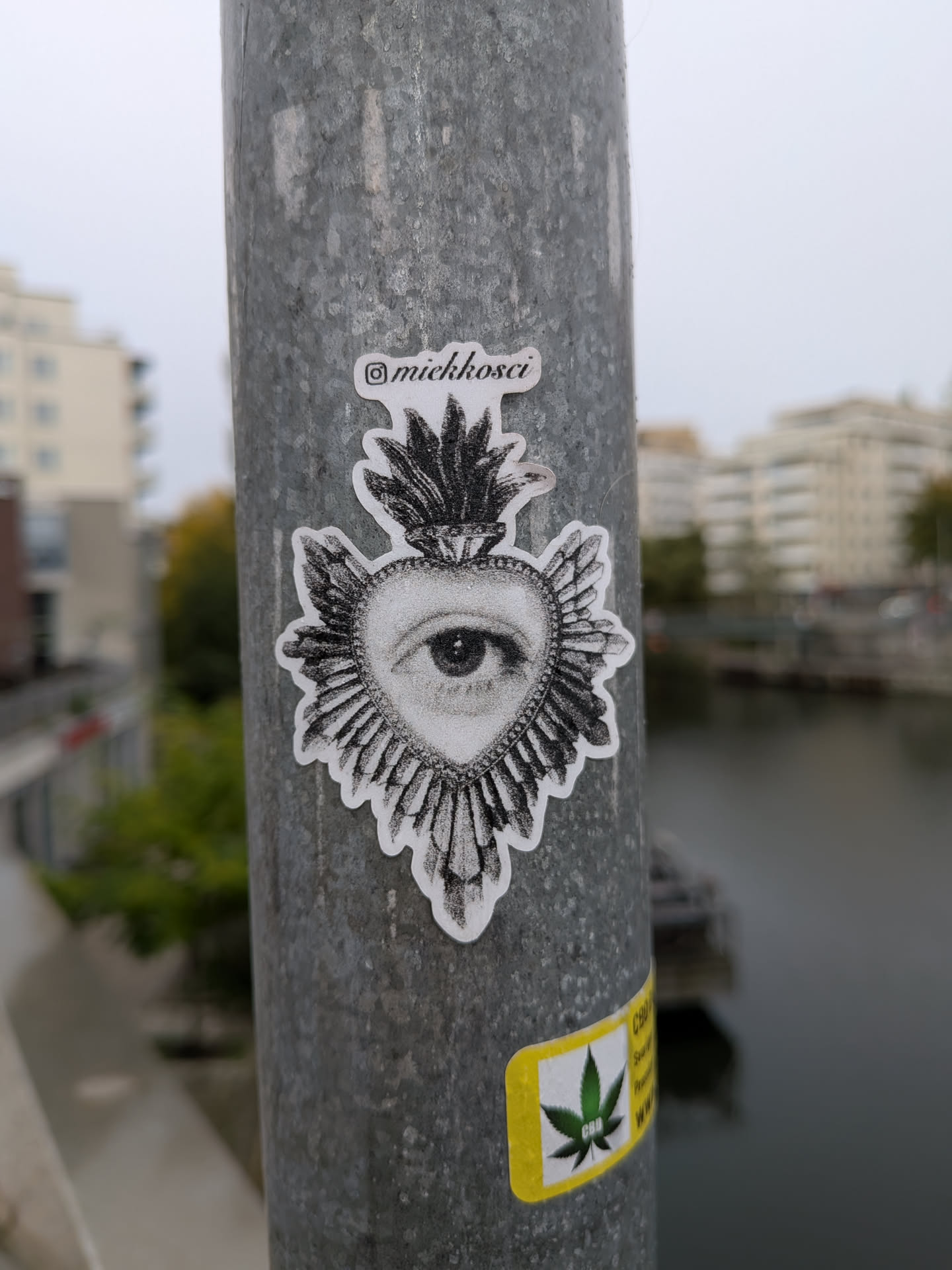 A die-cut black-and-white sticker placed vertically on a galvanized metal pole. The artwork features a sacred heart motif adorned with radiating spikes or rays and topped with flames. In the center of the heart is a realistically drawn human eye executed in a fine stippling and dotwork style. At the top of the sticker, an Instagram logo precedes the cursive handle '@miekkosci'.