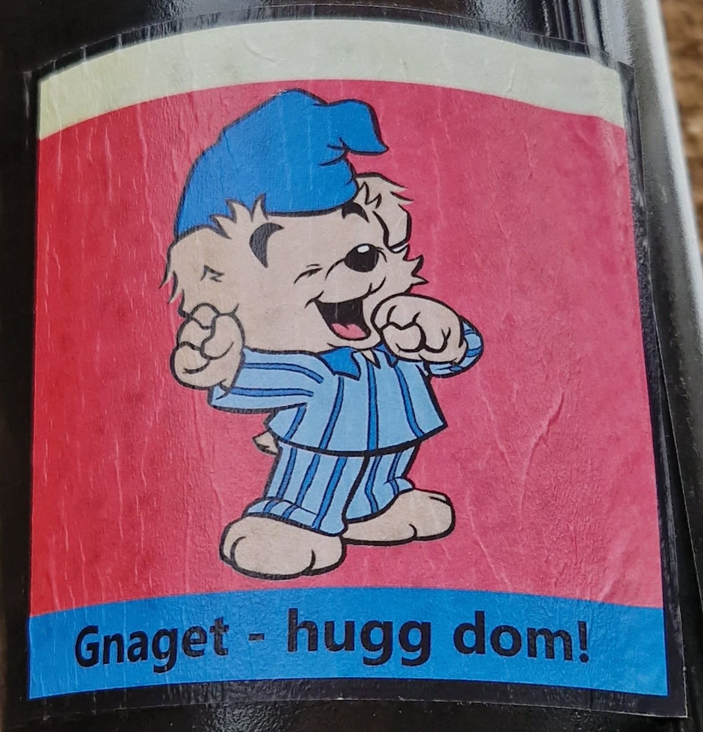 A sticker featuring a cartoon illustration of a bear wearing blue pajamas and a sleeping cap. The bear has a cheerful expression and is shown with its fists slightly clenched. The background is pink, and there is a blue banner at the bottom with the text: "Gnugget - hugg dom!"