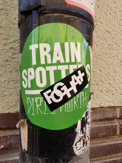 A green circular sticker with the text "TRAIN SPOTTERS" in white. On top of it, a smaller black sticker with the text "FOGH#" in white and illegible text underneath is applied. The bottom part of the main sticker has illegible text. The stickers are affixed to a black pole.
