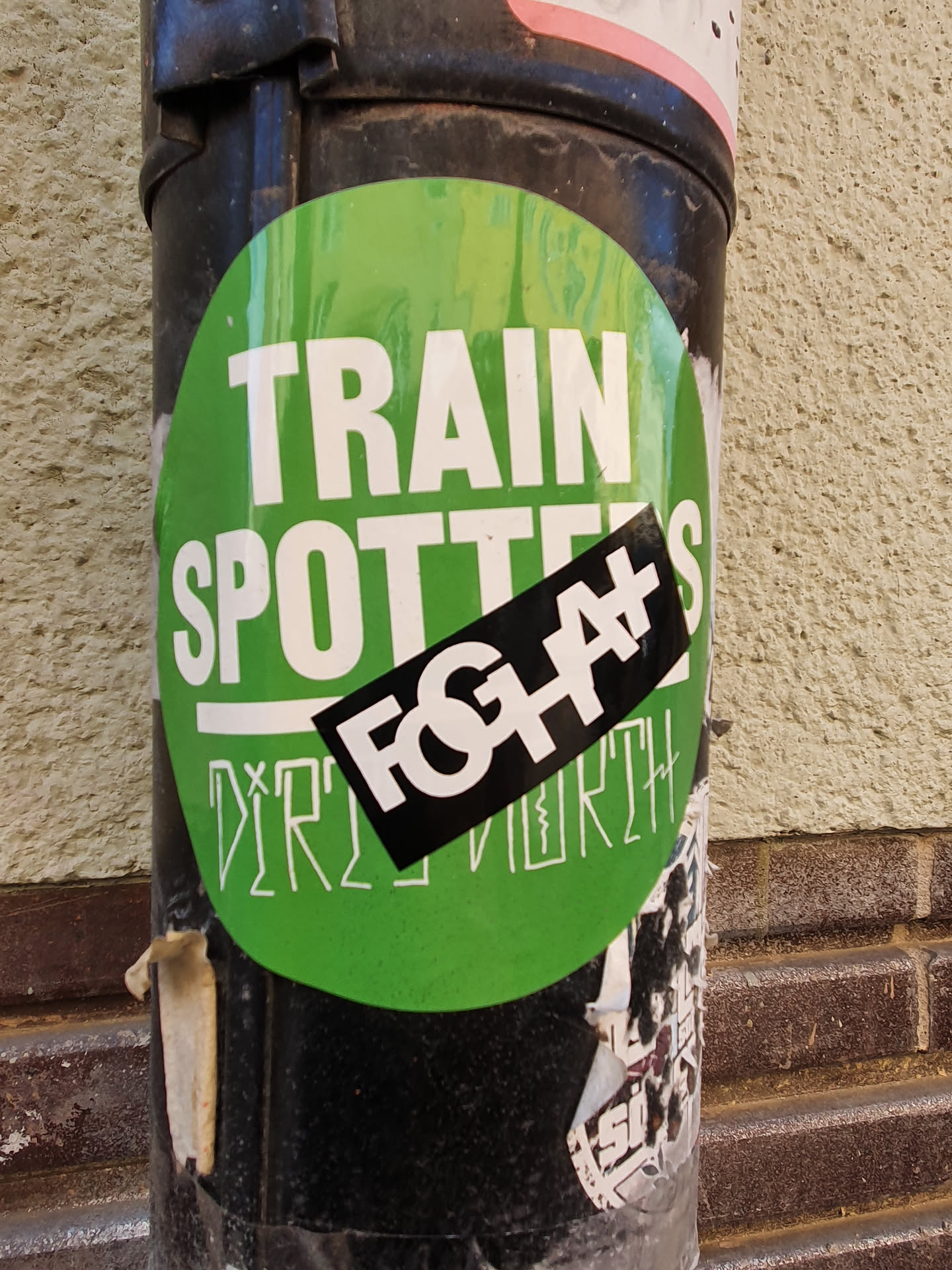 A green circular sticker with the text "TRAIN SPOTTERS" in white. On top of it, a smaller black sticker with the text "FOGH#" in white and illegible text underneath is applied. The bottom part of the main sticker has illegible text. The stickers are affixed to a black pole.