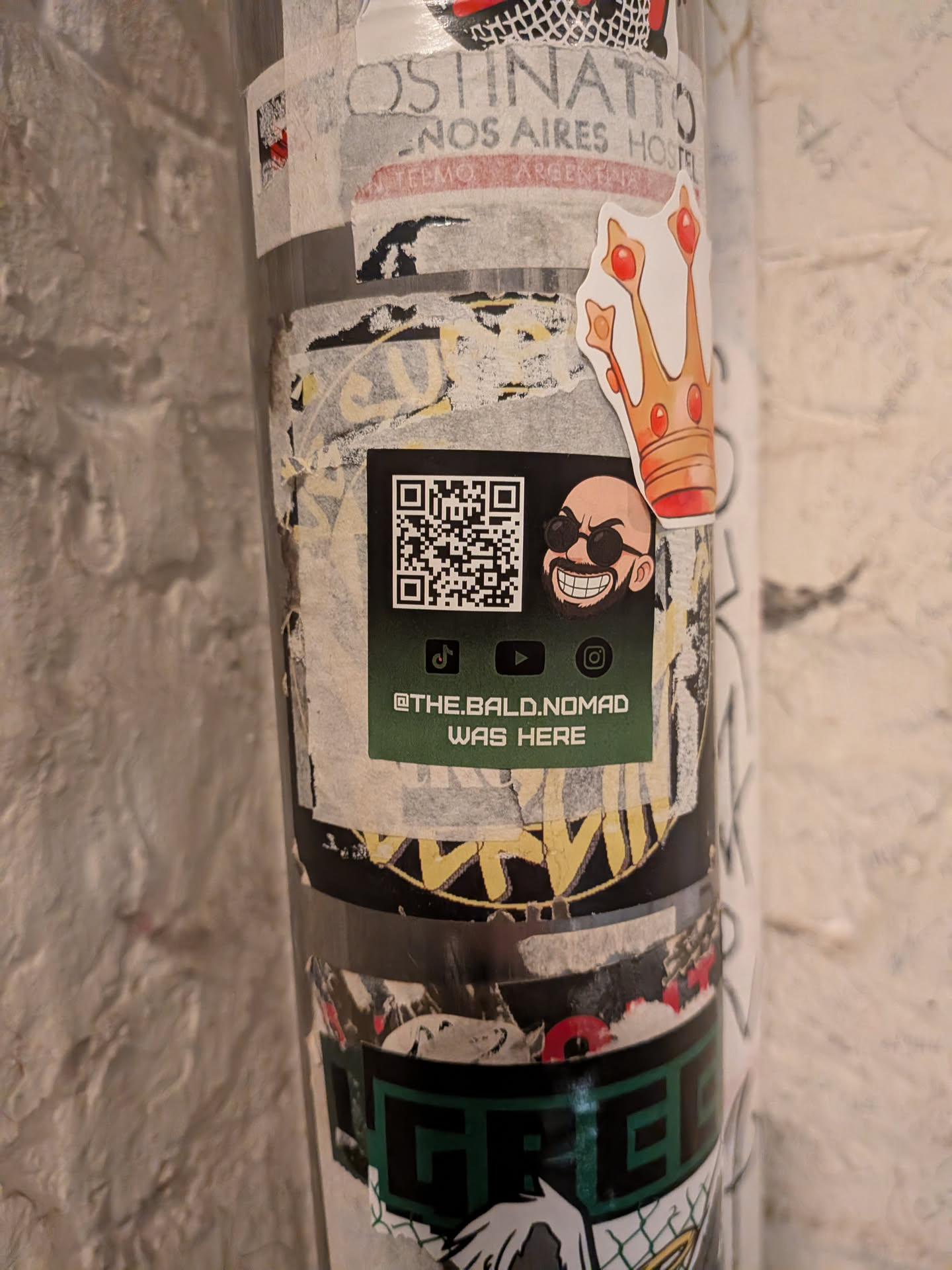 A dark green rectangular sticker affixed to a post over older, worn stickers. The upper section features a scannable QR code on the left and a cartoon illustration of a smiling bald man with a beard and dark sunglasses on the right. Below these are small black icons for TikTok, YouTube, and Instagram, followed by the white stylized text '@THE.BALD.NOMAD WAS HERE'.