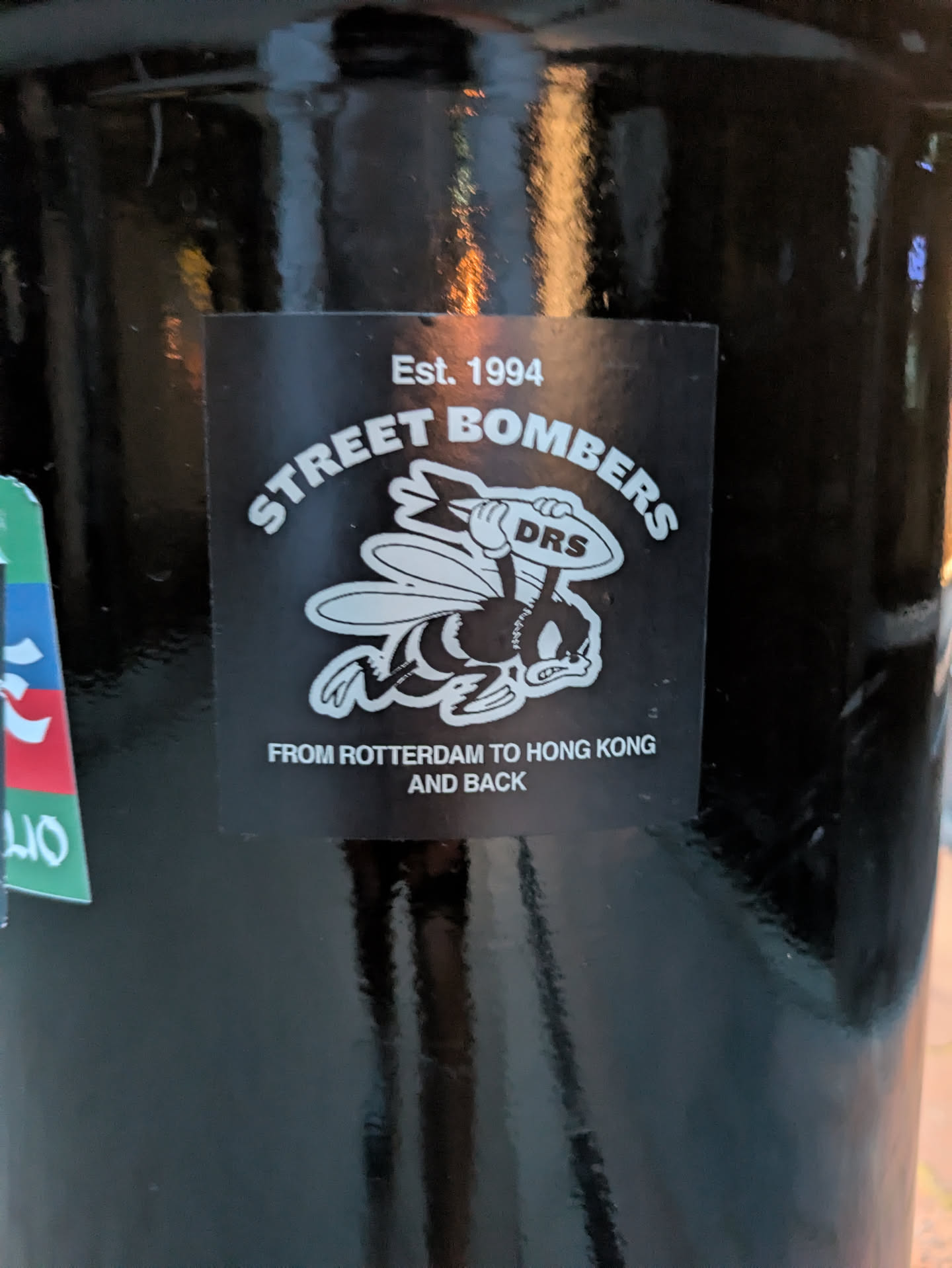 The sticker is square with a black background. At the top, it reads "Est. 1994" in white text. Below this, in a curved white font, is "STREET BOMBERS". Centered below this text is a cartoon illustration of a bee with wings outstretched, wearing boxing gloves, and carrying a rugby ball with the letters "DRS" on it. The bee has a determined expression. Underneath the bee, in white capital letters, is the text "FROM ROTTERDAM TO HONG KONG AND BACK". The sticker appears to be slightly weathered.