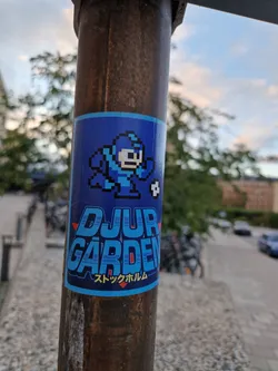 A blue sticker featuring pixel art of Mega Man from the classic video game series. The sticker is placed on a brown metal pole. The text on the sticker says "DJUR GARDEN" and "ストックホルム" (Stockholm in Japanese).
