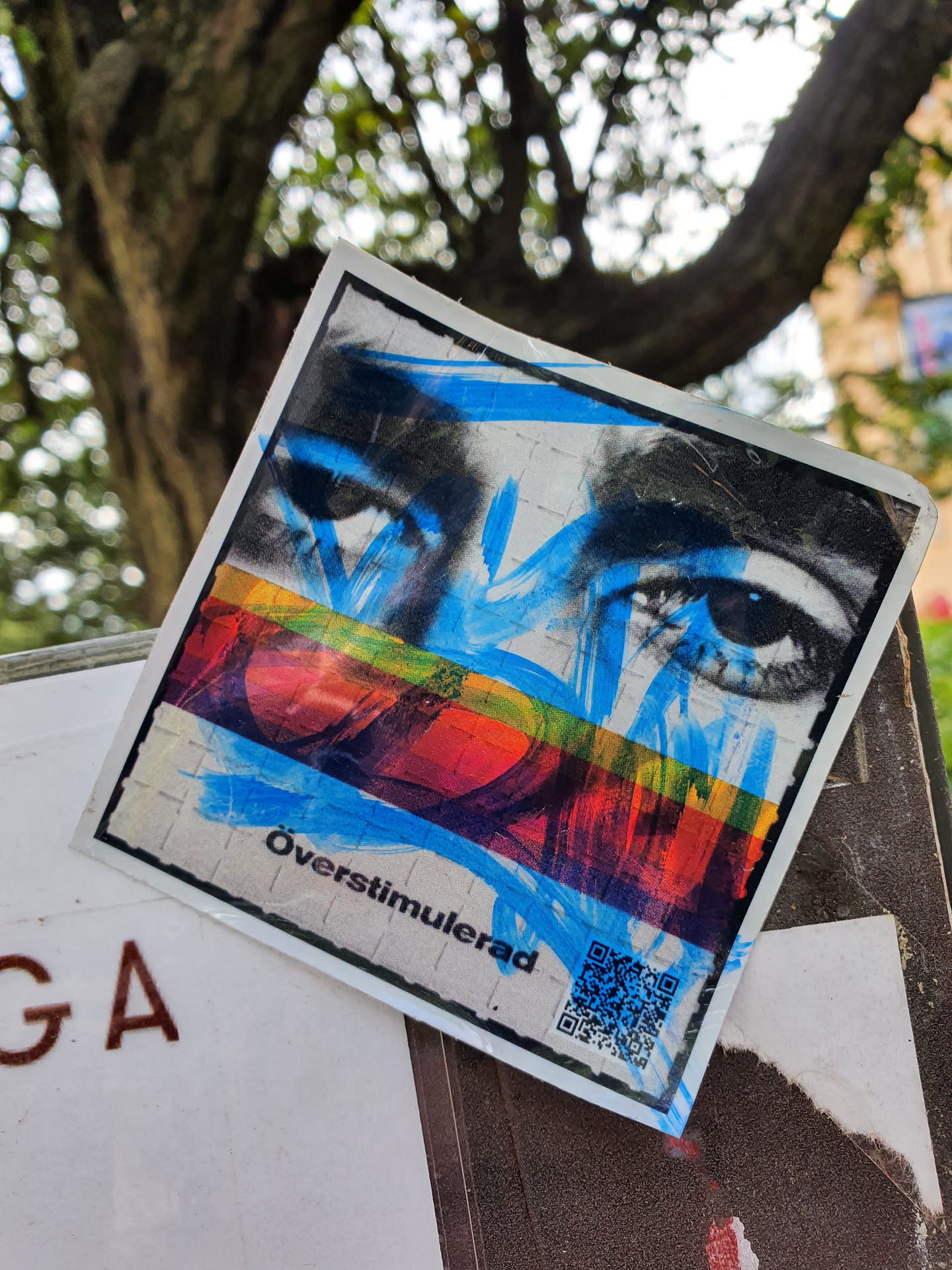 A sticker featuring a stylized image of eyes with blue paint strokes and a rainbow colored stripe.  The text 'Överstimulerad' is visible at the bottom.