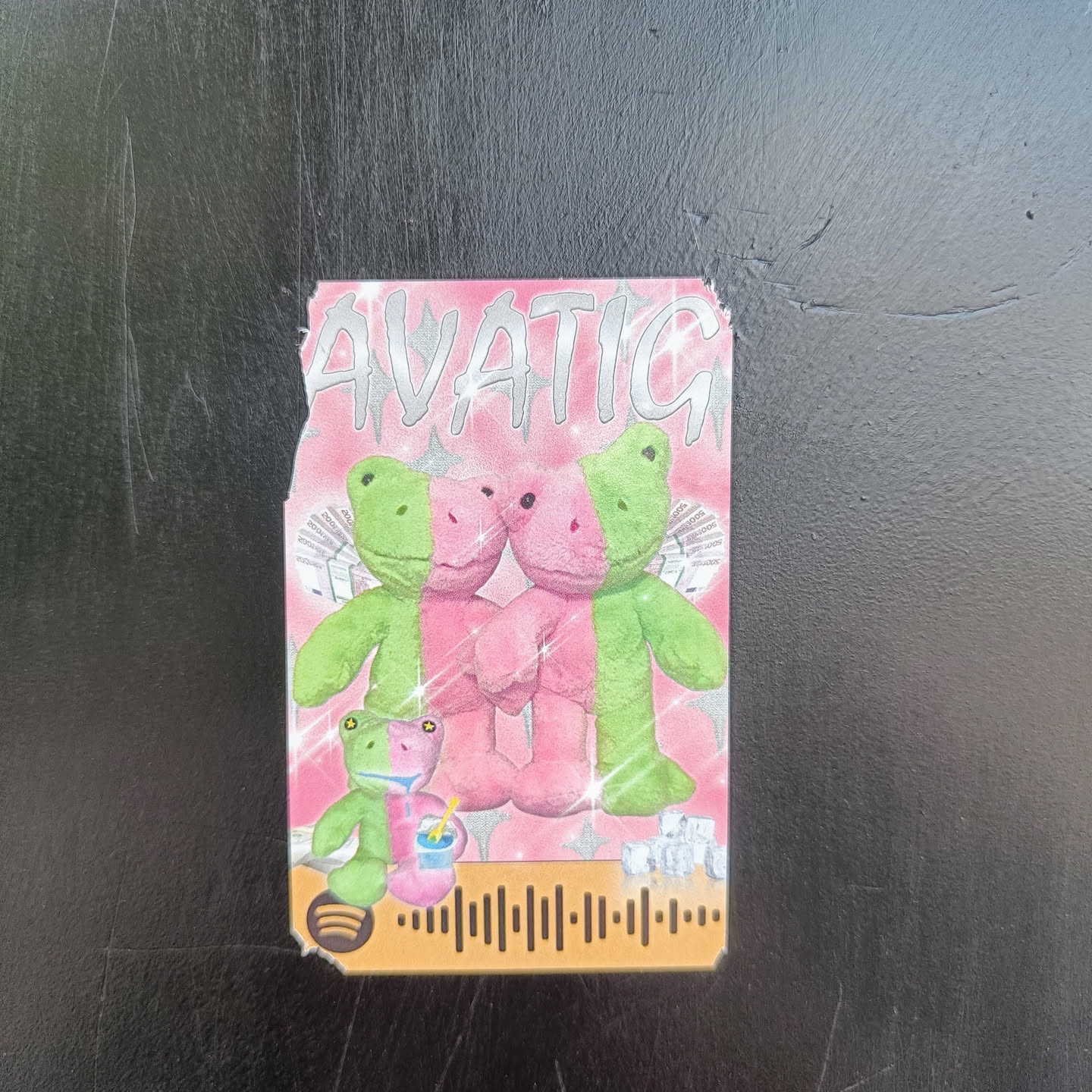 Pink and green frog plushies sticker with a Spotify code. The sticker features two frog plushies holding hands, with a smaller frog plushie holding a drink below them. The background is pink with sparkles and the word "AVATIC" written in a stylized font at the top. A Spotify code is visible at the bottom.
