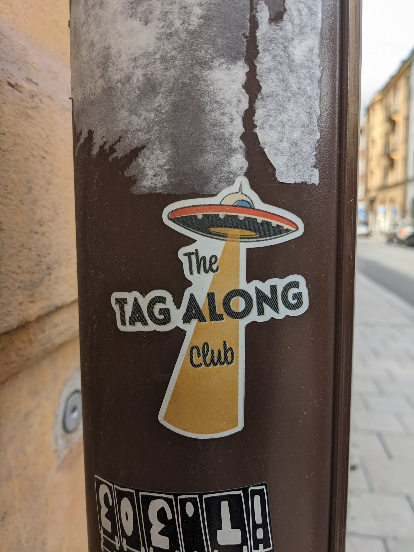 A sticker featuring an illustration of a UFO and the text "The Tag Along Club". The sticker has a retro style and is on a brown pole.