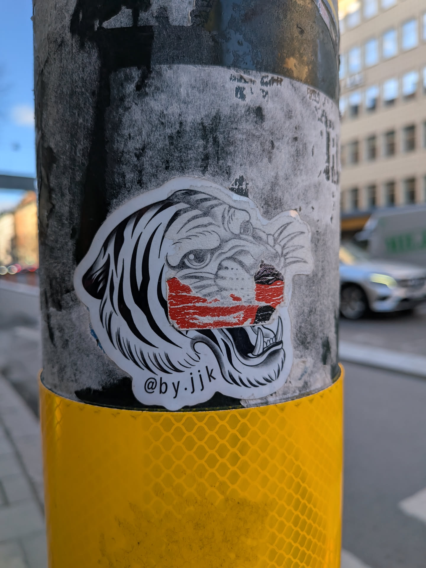 The sticker features a stylized illustration of a tiger's head, predominantly in black and white. The tiger has bold black stripes on a white background, with detailed eyes and an open mouth revealing fangs, conveying a fierce or roaring expression. A prominent, distressed red rectangular patch covers part of its face, specifically across the nose and upper jaw area, creating a striking contrast and a 'censored' or 'damaged' aesthetic. The sticker has a clean white border, cut out around the contour of the tiger's head, and is applied to a dark, worn pole.