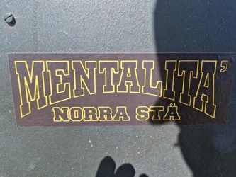 A dark brown sticker with yellow text. The text says "MENTALITÄ" in a large, bold font, with "NORRA STÄD" written underneath in a smaller font.