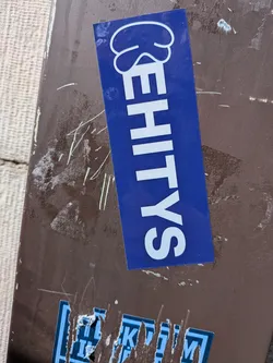 A rectangular sticker with a vibrant blue background, oriented vertically. The word "EHITYS" is written in bold, sans-serif, uppercase white letters, stacked from bottom to top. Above the 'E' (at the top of the sticker), there is a white, stylized graphic resembling a cloud or a thought bubble. The sticker has a glossy finish and is applied to a dark brown, textured surface.