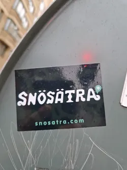 A black sticker with the word Snösätra written in a stylized font.  The website snosatra.com is printed underneath. A small snowflake icon is located to the right of the text.