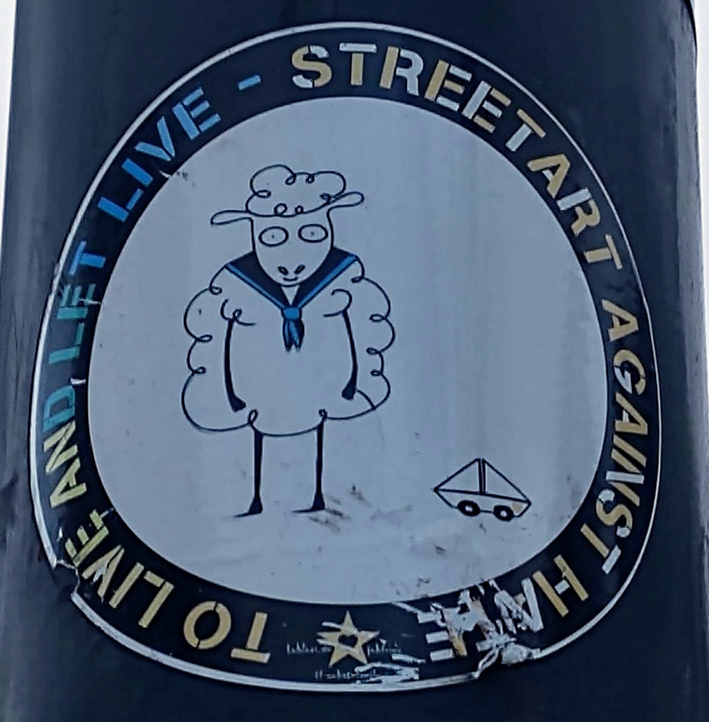 Round sticker featuring a cartoon sheep wearing a sailor's outfit.  A small boat is depicted below the sheep. The text "To Live And Let Live - Street Art Against The Machine" circles the illustration.