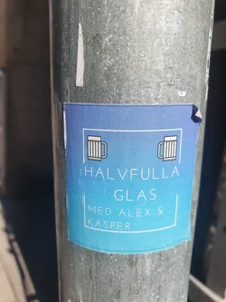 A blue sticker with a simple design featuring two beer mugs and the text 'HALVFULLA GLAS MED ALEX & KASPER'.